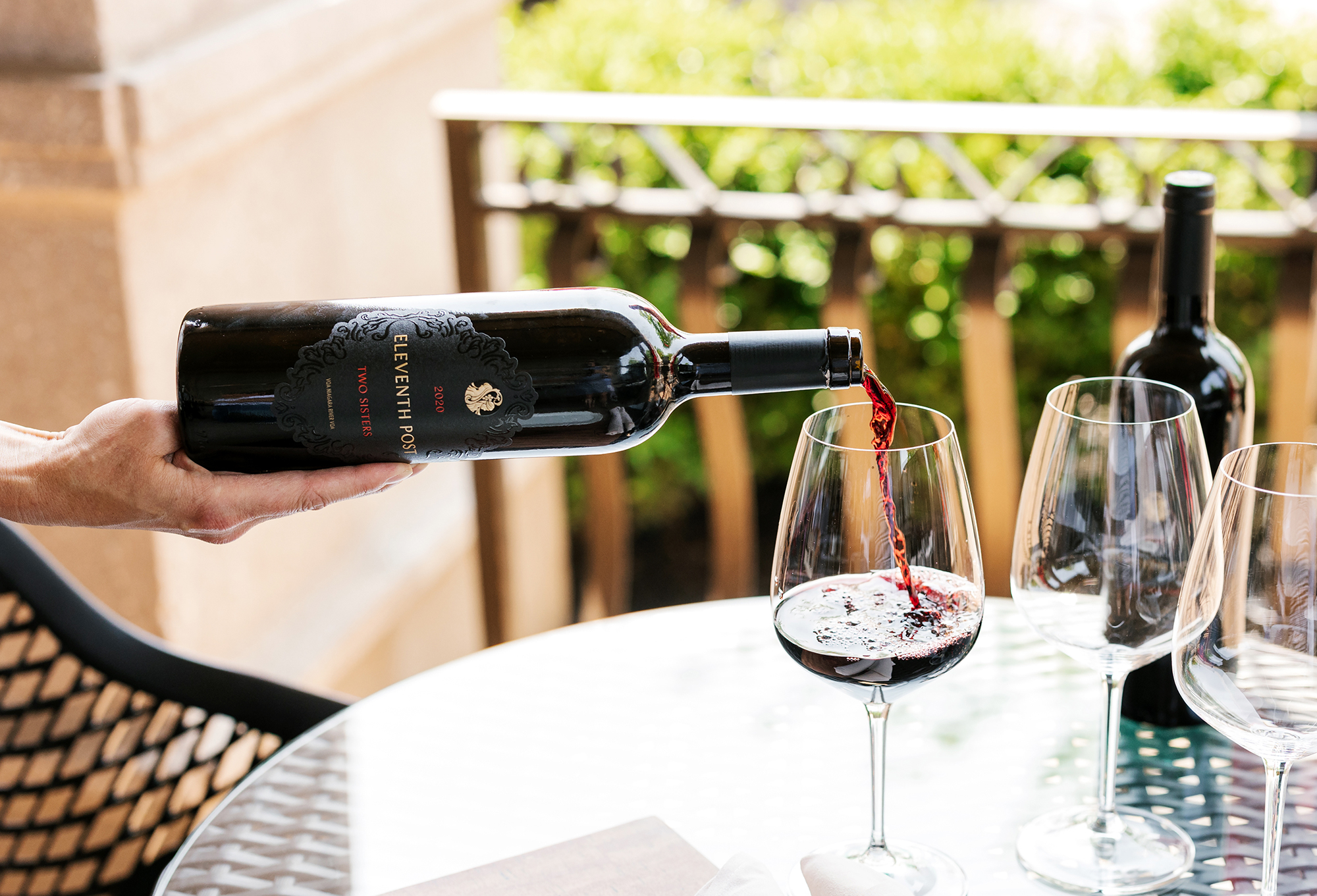 Red wine from a Two Sisters Vineyards bottle being poured into a glass on an outdoor table, with additional wine glasses and a bottle in the background
