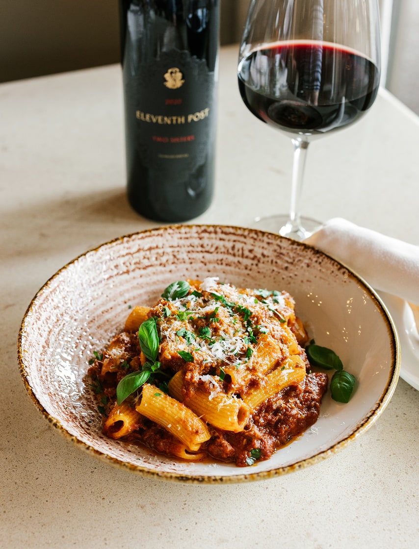 Bowl of rigatoni with meat sauce and basil served alongside a glass of red wine and a bottle of 2020 Eleventh Post by Two Sisters Vineyards