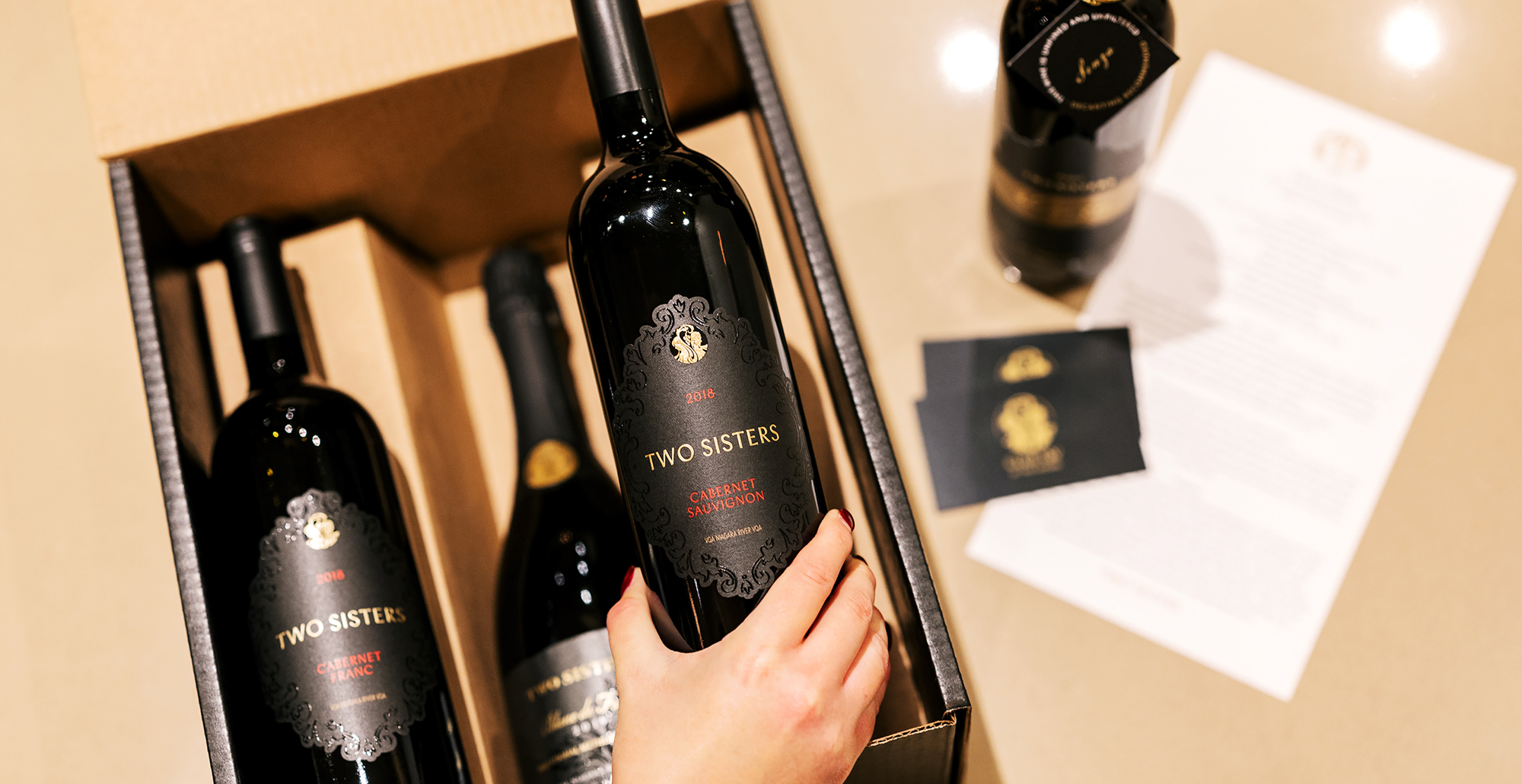Hand placing a bottle of Two Sisters Cabernet Sauvignon into a wine gift box with additional bottles and club materials