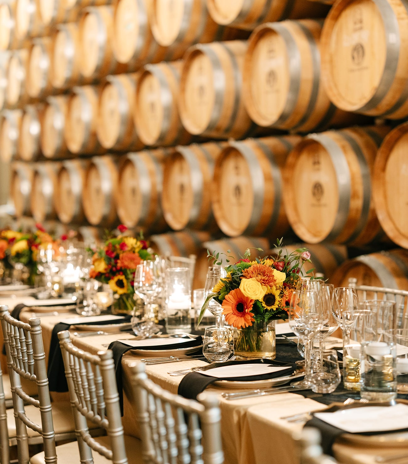 Elegant winery event setup with banquet tables decorated with floral centerpieces, wine glasses, and candles inside a barrel room