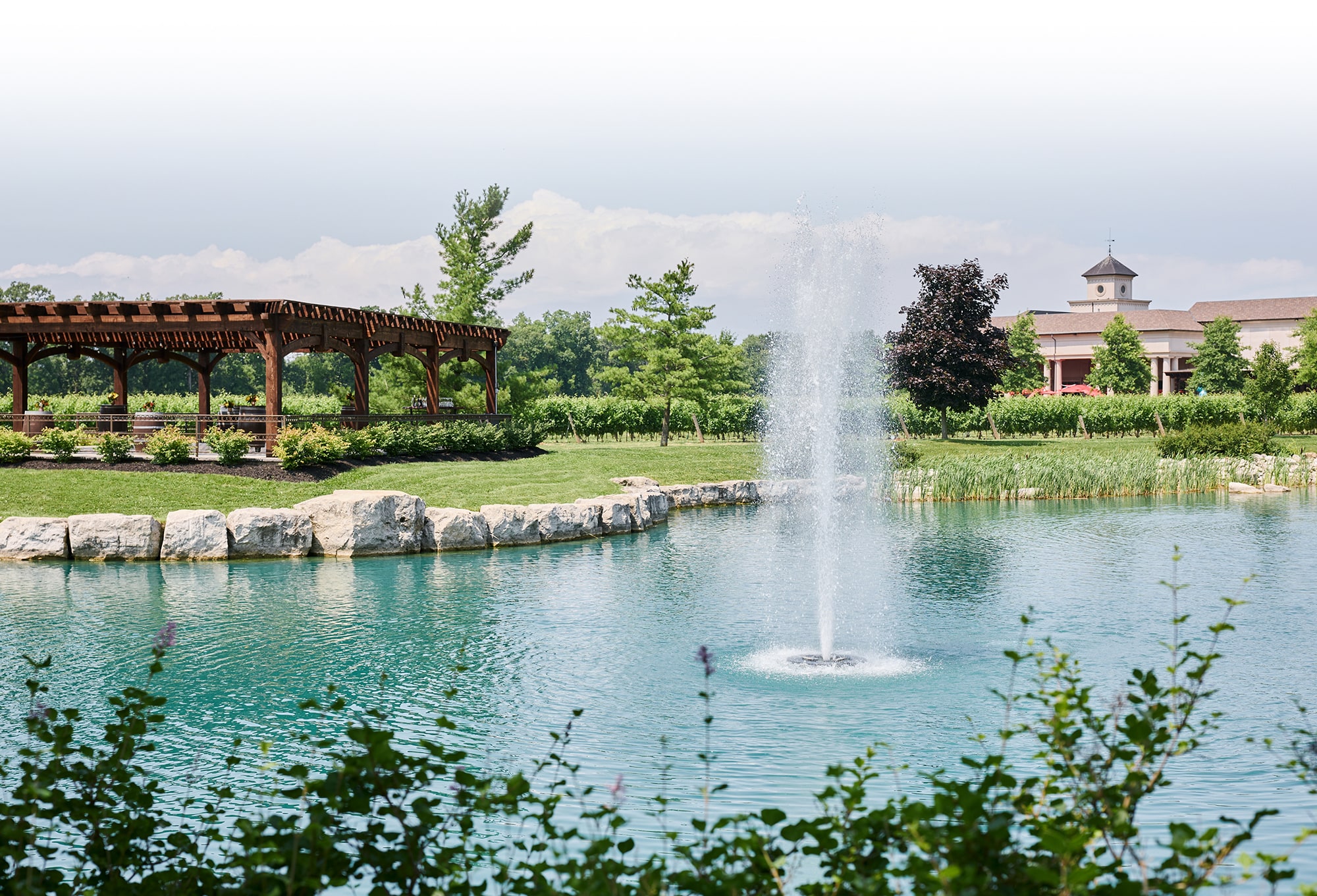 Scenic pond with a central fountain at a landscaped estate, surrounded by greenery, a wooden pergola, and a vineyard-style building in the background