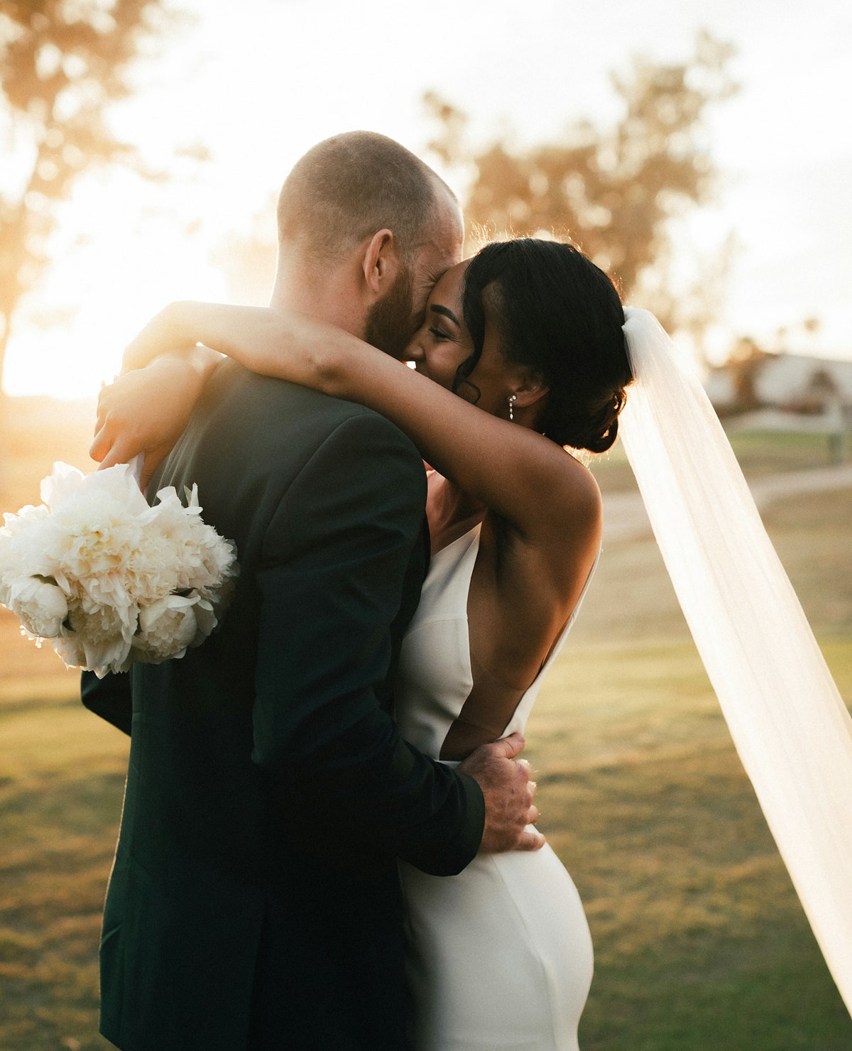 Bride and groom embrace at sunset on a grassy outdoor venue, the bride holding a bouquet and wearing a long veil flowing behind her