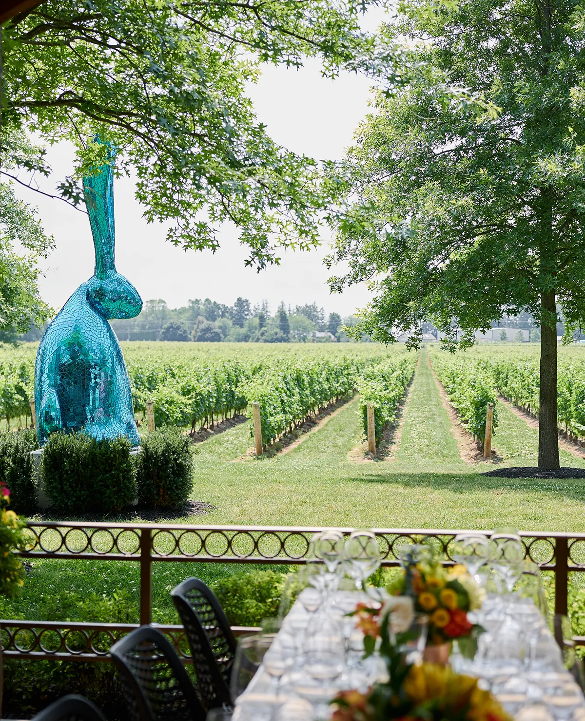 Outdoor winery dining table set with wine glasses, floral center pieces, blue bunny sculpture and vineyard view at Two Sisters Vineyards Niagara.